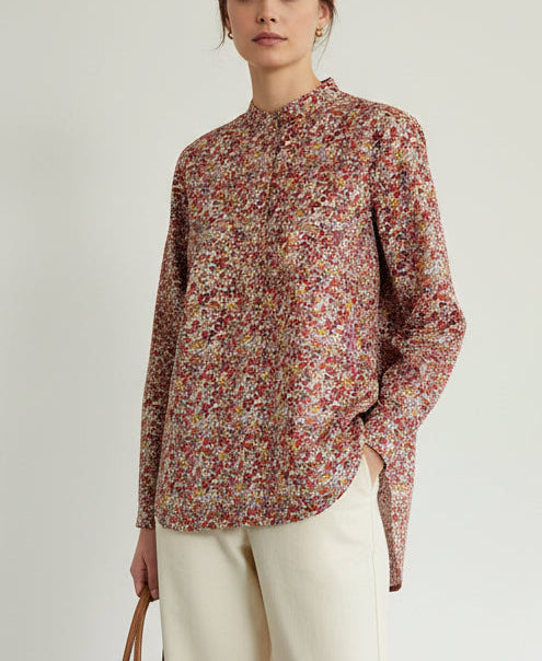 A long-sleeve button-up shirt with a dense red, pink, and yellow floral print, hanging on a wooden hanger against a plain off-white wall.