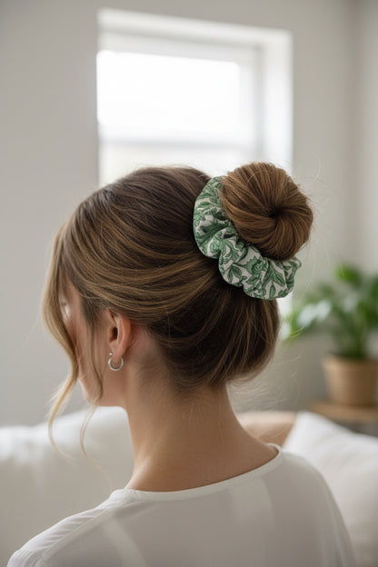A fabric scrunchie with a green botanical leaf pattern on a white background, photographed flat against a light off-white surface.